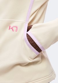 Main dans la poche d'un vêtement à manches longues couleur crème avec bordure violet clair et petit logo rose brodé près de la poche.