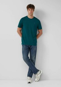 Junger Mann, der gegen eine Wand steht und ein türkisfarbenes T-Shirt mit kurzen Ärmeln, blue Jeans und weiße Sneakers mit grünen Akzenten trägt.