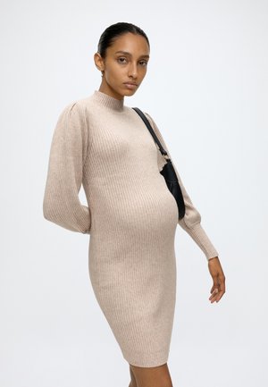 Mujer embarazada con vestido de punto beige con canalé y bolso negro de hombro sobre un fondo blanco liso.