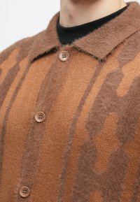 Καφέ κασμίρηνο πουλόβερ με υφή, διαθέτει γιακά, πέντε κουμπιά και γεωμετρικό σχέδιο σε πιο ανοιχτές τόνους.