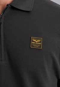 Donkergrijze polo met een ritskraag, voorzien van een vierkant zwart en gouden metalen logo-opdruk op de linkerkant van de borst. Gelaagd stof.