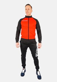 Rdeča in črna trenirka z zip-jakom in usklajenimi hlačami. Nosil logo na jakni in ob strani hlač. Vključuje superge.