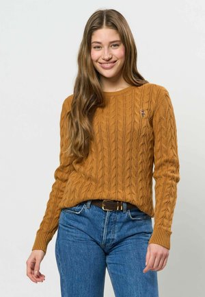Jeune femme aux longs cheveux bruns, portant un pull en maille torsadée moutarde et un jean bleu, souriant sur un fond clair uni.