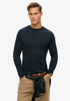 Chemise à manches longues bleu marine avec un col rond, ornée d'un logo discret. Associée à un pantalon kaki et une chemise à carreaux verte nouée autour de la taille.