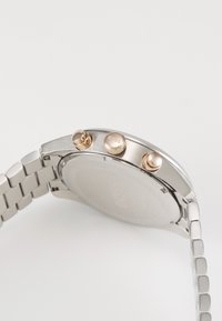 Orologio in acciaio inox argento con cinturino texturizzato, tre pulsanti color oro e cassa lucida. Il retro è liscio con un'impronta del logo.