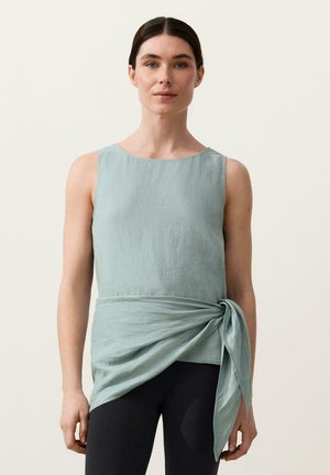 Sötét hajú nő világoskék ujjatlan felsőben, melynek derékpántja van, fekete leggingssel, egyszerű, világos háttér előtt állva.