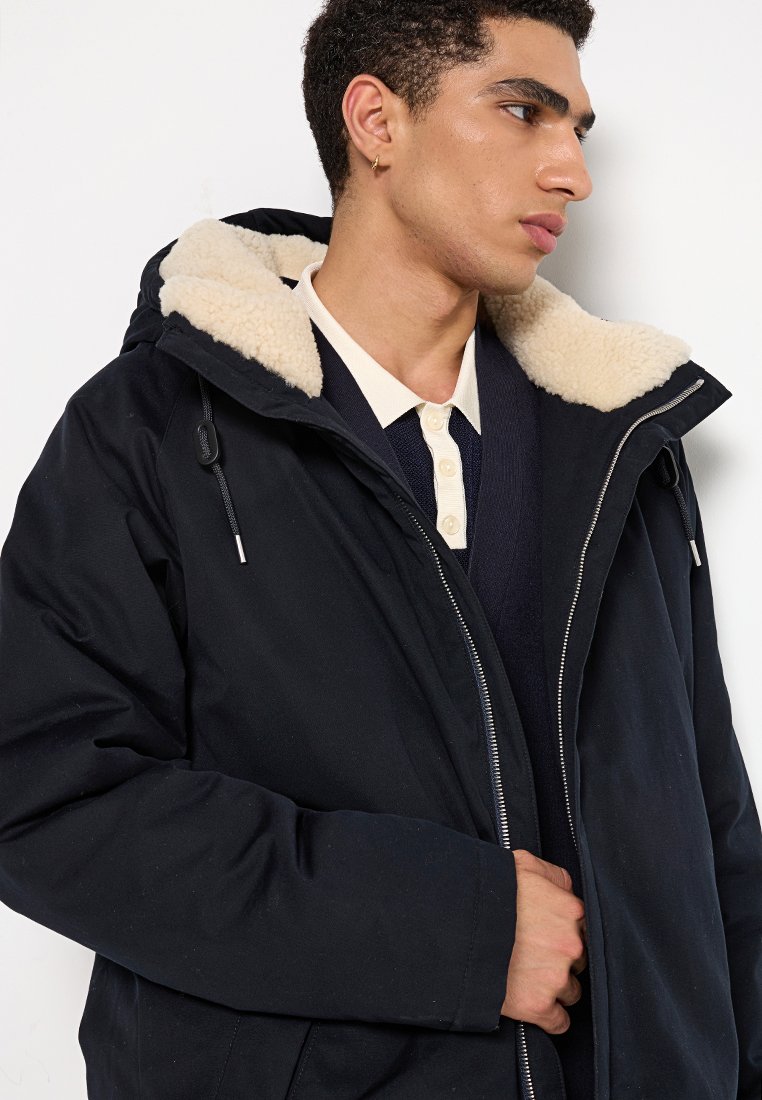 Marineblauwe geïsoleerde jas met een faux fur gevoerde capuchon, voorrits en trekkoord. Draagt over een donkere trui met een lichtgekleurde kraag.