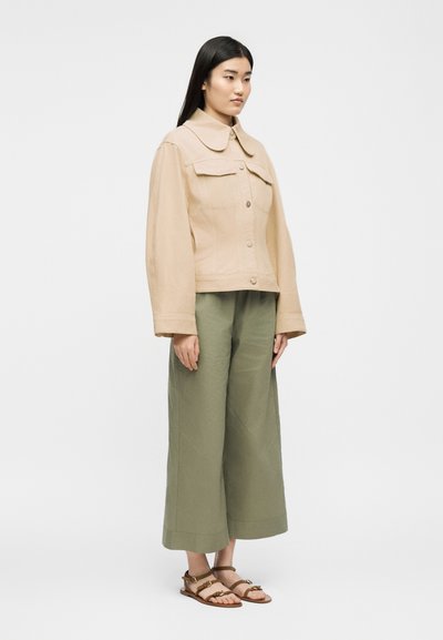 Γυναίκα που στέκεται, φοράει μπεζ σακάκι με κουμπιά, φαρδύ παντελόνι cropped σε ελιά απόχρωση και ταμπά φλατ σανδάλια, με φόντο απλό λευκό.