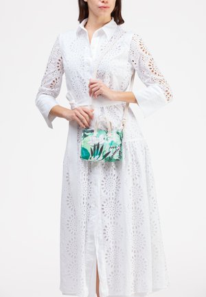 Borsa a mano in plastica trasparente con motivo floreale verde e bianco, dotata di due manici superiori e di una tracolla staccabile, è tenuta davanti a un vestito bianco ricamato.