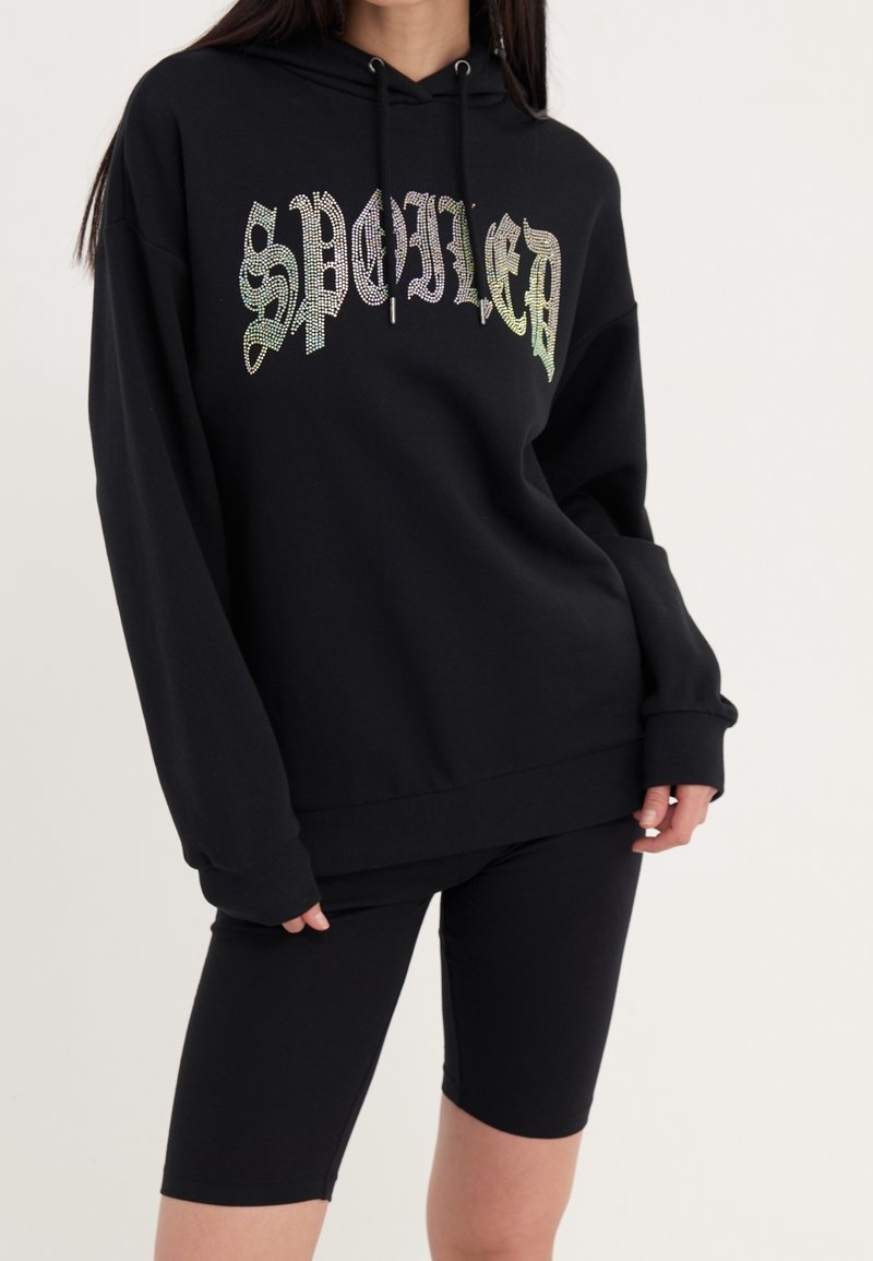 Felpa nera con la scritta "SPOILED" in strass iridescenti. Il materiale è morbido con una vestibilità rilassata, abbinata a pantaloni corti neri.