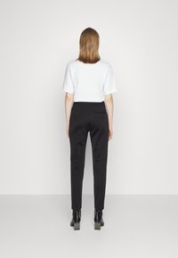 Černé ušité kalhoty s hladkou texturou a úzkým střihem, v kombinaci s jednoduchou bílou košilí s krátkým rukávem, pohled zezadu.