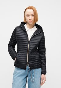 Γυναίκα με ίσια κόκκινα μαλλιά φοράει μαύρο μπουφάν με καπιτονέ πάνω από λευκό πουκάμισο με γιακά και ανοιχτόχρωμο τζιν, στέκεται μπροστά σε απλό φόντο.