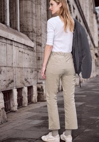 Femme aux cheveux longs portant un pantalon beige, un haut blanc et des baskets, tenant une veste grise sur son épaule, debout sur un trottoir en ville.
