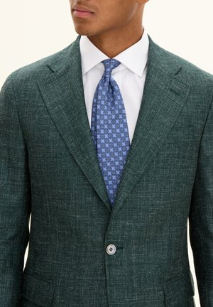 Uomo che indossa un blazer testurizzato verde scuro, una camicia bianca e una cravatta blu a fantasia, abbottonato sul davanti.