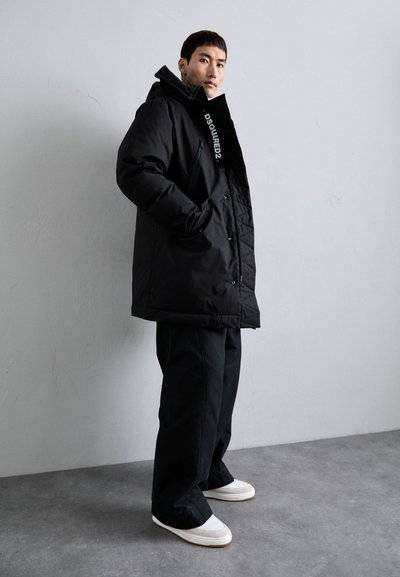Black padded parka with a hood, featuring a branded zip and button closure, oversized fit, and side pockets, paired with black trousers.