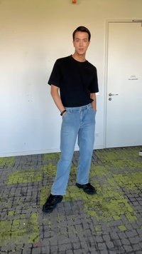 Νέος άνθρωπος στέκεται μέσα σε εσωτερικό χώρο δίπλα σε λευκό τοίχο και κλειστή πόρτα, φοράει μαύρο πουκάμισο, ανοιχτόχρωμο τζιν και μαύρα παπούτσια, με τα χέρια στις τσέπες.