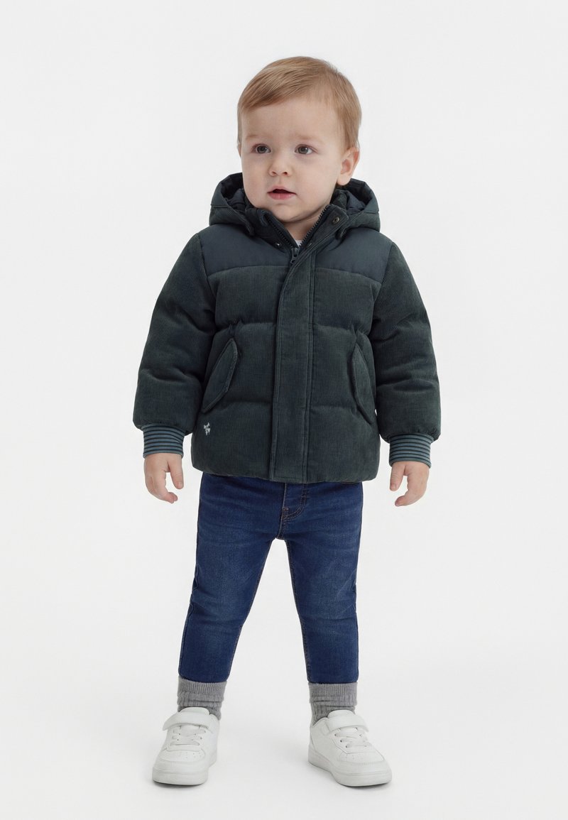 Tout-petit debout portant une doudoune à capuche vert foncé, un jean bleu, des chaussettes grises et des baskets blanches sur un fond blanc uni.