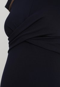 Mörkblå klänning med en figurnära design och tvistad tygdetalj i midjan, tillverkad av ett mjukt, elastiskt material. Slät textur.