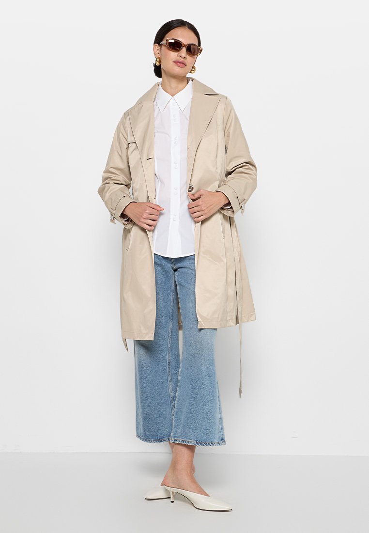 Trench-coat beige avec taille cintrée, tissu léger, manches longues et col à cran, porté sur une chemise blanche et un pantalon en denim évasé.