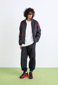 Veste de survêtement noire avec accents rouges, associée à un pantalon noir assorti et des baskets avec accents rouges. Le modèle se tient sur une surface verte.