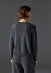 Temno siva pletena jopica z raglan rokavi, rebrastim ovratnikom in robom. Mehka tekstura, enostaven dizajn, nošena z usklajenimi temno sivimi hlačami.