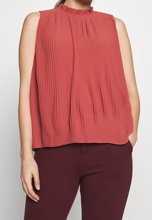 Blouse sans manches plissée rouge rouille avec col à volants portée avec un pantalon bordeaux foncé, montrant le torse et les bras.