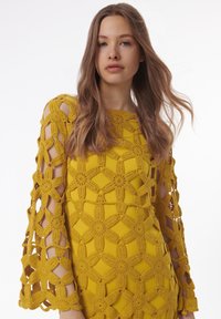 Top en crochet jaune présentant un motif floral avec des détails en dentelle ouverte, des manches longues et un col arrondi. Apparence texturée.