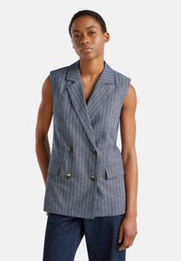 Dvojřadý bezrukávový blazer z modré látky s bílými pruhy, se dvěma předními kapsami a zlatými knoflíky. Přiléhavý střih.
