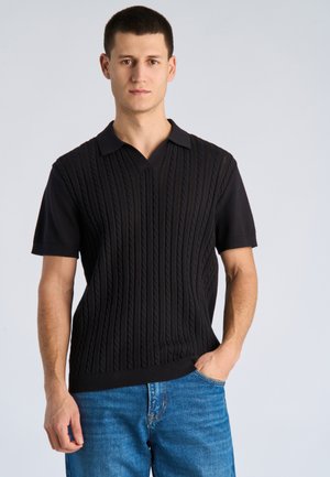 Jonge man staand met hand in zijn zak, draagt een zwarte gebreide polo met korte mouwen en een blauwe spijkerbroek, tegen een effen lichte achtergrond.