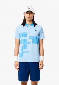Ljusblå polo med geometriska blå mönster, kombinerad med marinställda shorts. Outfiten inkluderar en vit basebollkeps och ett armband.