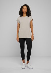 Ljusgrå kortärmad t-shirt med avslappnad passform, parat med svarta leggings och vita sneakers, mot en enfärgad grå bakgrund.