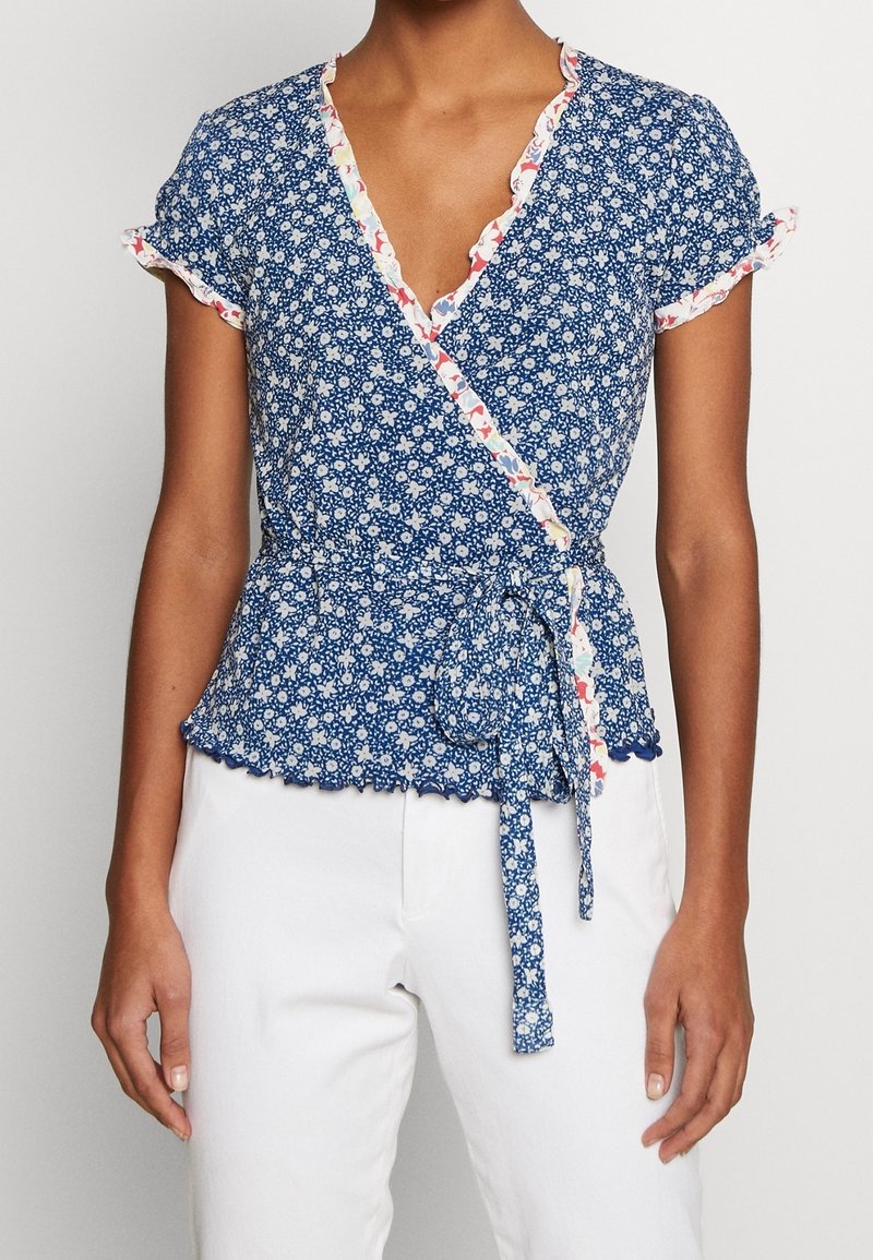 Femme portant un chemisier cache-cœur bleu à motif floral avec des bordures à volants et un pantalon blanc, debout devant un fond clair uni.