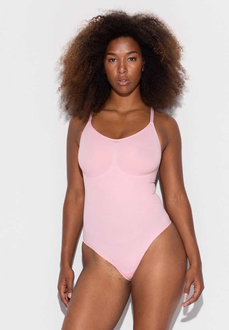 Femme aux cheveux afro portant un body sans manches rose clair, debout devant un fond blanc uni.