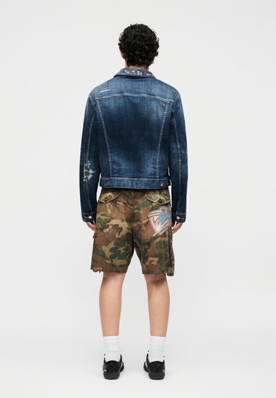 Άτομο με σγουρά μαλλιά στέκεται με την πλάτη, φορώντας ένα φθαρμένο μπλε τζιν μπουφάν, καμουφλάζ cargo σορτς με γραφικό ετικέτα, λευκές κάλτσες και μαύρα παπούτσια.