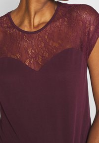 Burgundské tričko s krátkým rukávem, které má krajkový přehoz na výstřihu se zvlněným designem; hladká látka v dolní části s volným střihem.