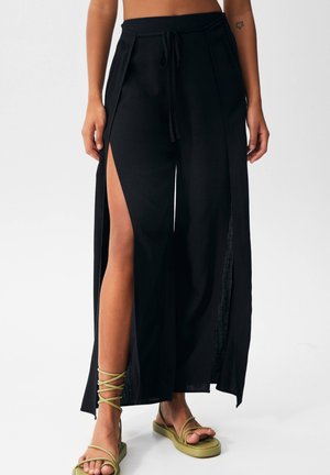 Frau trägt schwarze weite Hosen mit hohen seitlichen Schlitzen und olivgrüne flache Sandalen mit Riemchen, steht vor einem schlichten Hintergrund.