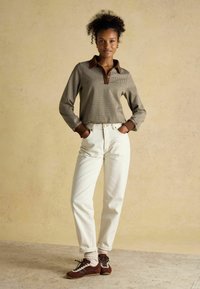 Brown and beige checked long-sleeve polo shirt, white high-waisted jeans, and brown shoes with laces. Model poses with hands in pockets.