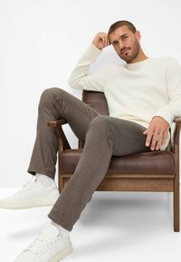 Graue strukturierte Hose kombiniert mit einem cremefarbenen gerippten Pullover und weißen Sneakers, sitzt auf einem Holzstuhl mit einem braunen Lederkissen.