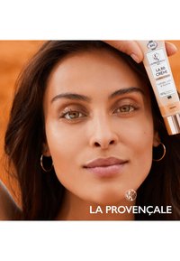 Tube de crème BB dans un emballage pêche clair avec un distributeur pompe, affichant la marque et les informations sur le produit, tenu près du visage d'un modèle.