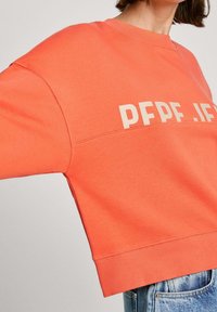 Korálová mikina s kulatým výstřihem, spadlými rameny a béžovým nápisem "PFPE.IF" na přední straně. Měkká tkanina s žebrovanými lemy.