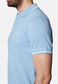 Jasnoniebieska koszulka polo z teksturowanej tkaniny, z trzema guzikami, oraz mankietami w białe paski. Posiada małe haftowane logo na piersi.