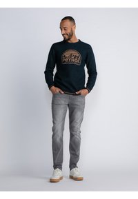 Navy Pullover mit einem braunen "PETROL"-Grafik in der Mitte, kombiniert mit hellgrauen Jeans und weißen Sneakers mit braunen Akzenten.