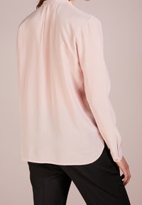 Blouse à manches longues rose clair avec une texture lisse, des poignets à boutons et un ourlet arrondi ; présente un pli subtil à l'arrière.