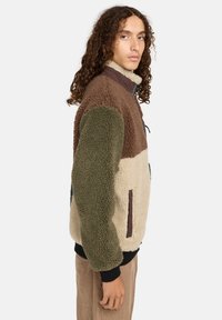 Veste en polaire avec un mélange de couleurs marron, vert et crème, dotée d'un col montant, d'une fermeture éclair et d'un tissu texturé.