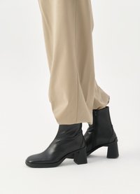 Cizme negre din piele, cu glezna, cu vârful pătrat și toc pătrat. Textură netedă, cu fermoar lateral. Asortate cu pantaloni bej, largi.
