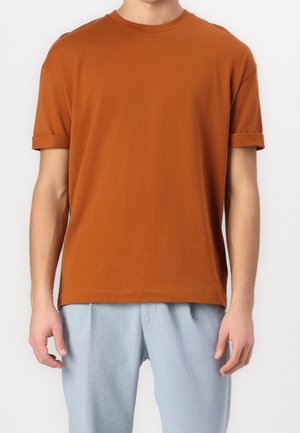 Adulto che indossa una t-shirt arancione scuro a maniche corte con risvolti e pantaloni azzurri chiari su sfondo bianco.
