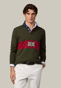 Zielona koszulka polo z długim rękawem, granatowym kołnierzem, czerwoną poziomą linią i emblematem Union Jack na piersi. Noszona z jasnymi spodniami i paskiem.