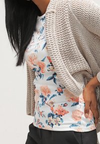 Kobieta z długimi ciemnymi włosami nosi beżowy dziergany kardigan na białej koszuli w kwiaty i czarnych spodniach, stojąc na jednolitym tle.