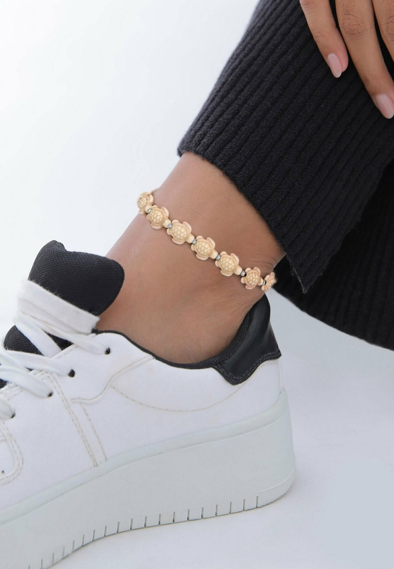 Guldtonet anklet med teksturerede blomstermotiver og små perler, båret sammen med en hvid sneaker med sort detalje.