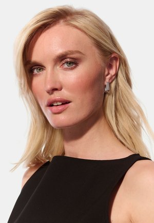 Nærbillede af en person med langt, glat blond hår, der bærer sølv hoopsøreringe og en sort ærmeløs top mod en lys baggrund.